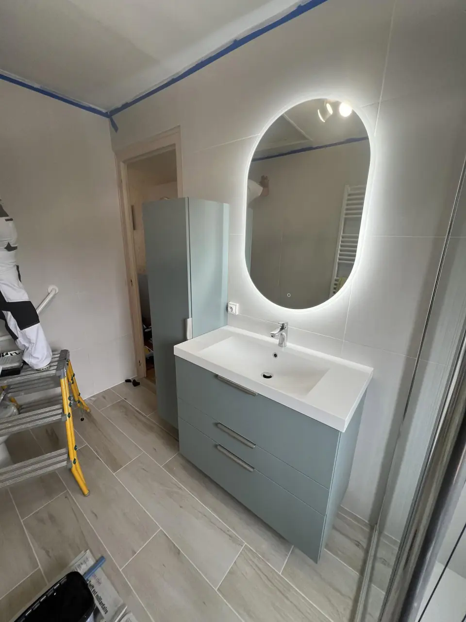 Salle de bains en finition : meuble vasque bleu-gris à tiroirs, miroir ovale rétroéclairé et colonne de rangement assortie