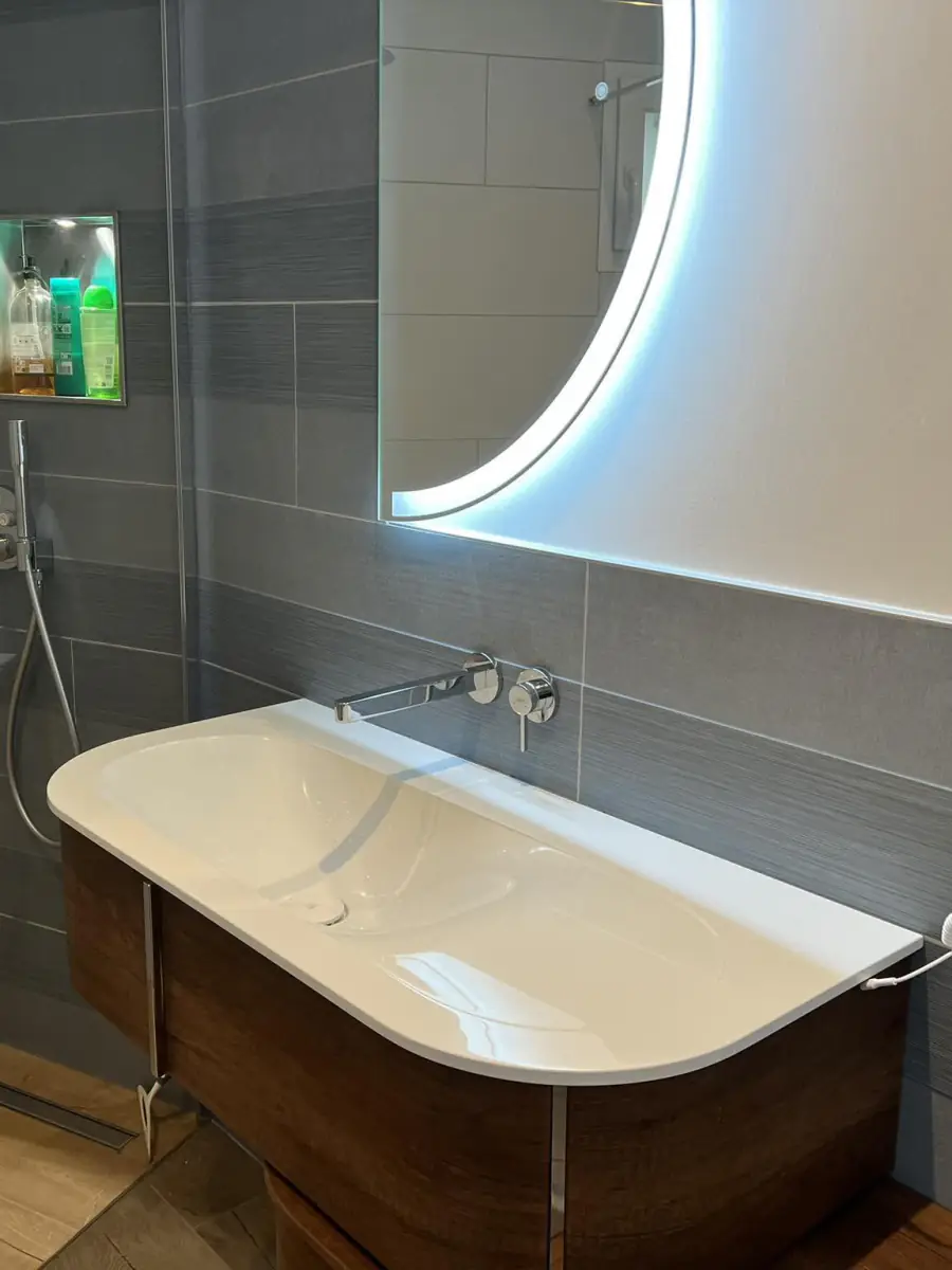 Salle de bains rénovée : vasque blanche sur meuble suspendu en bois foncé, robinetterie murale chromée, miroir rond rétroéclairé et douche à l’italienne